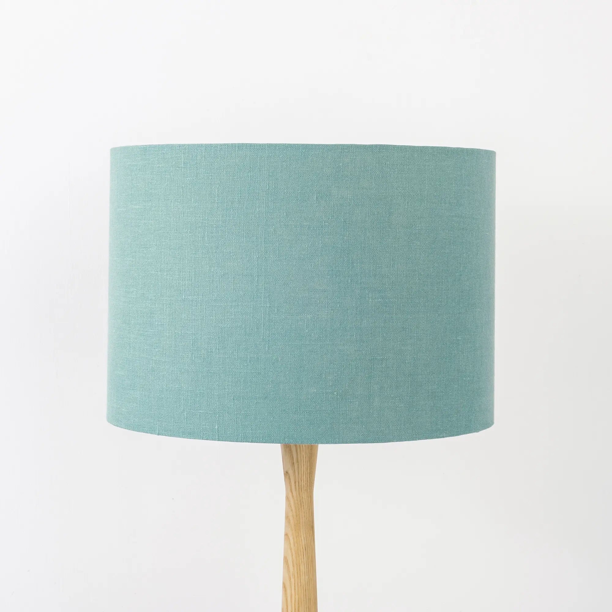 A dusty turquoise linen lampshade on a wooden lamp base, with a textured weave design, photographed against a white background.