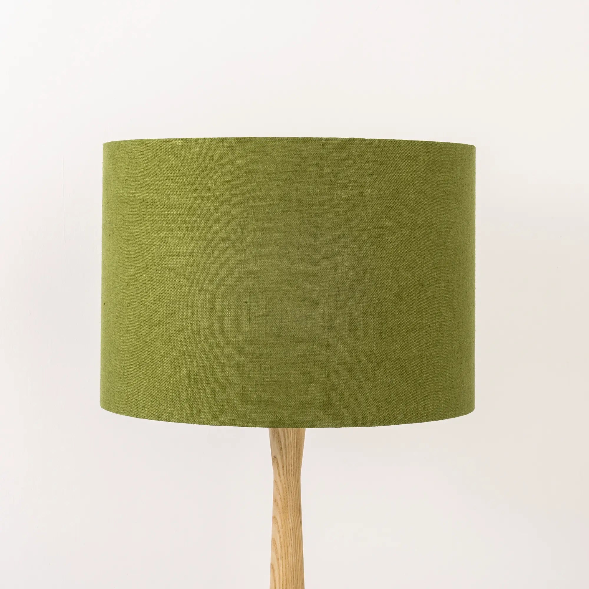 A moss green linen lampshade on a wooden lamp base, with a textured weave design, photographed against a white background.