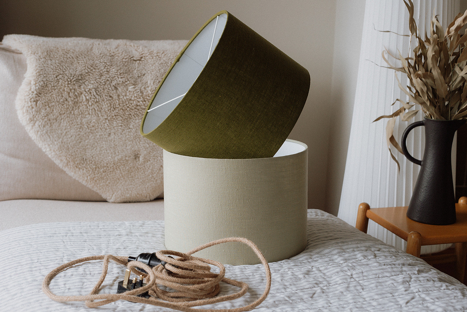 Two green drum lampshades sitting on a bed next to a vase of dried flowers